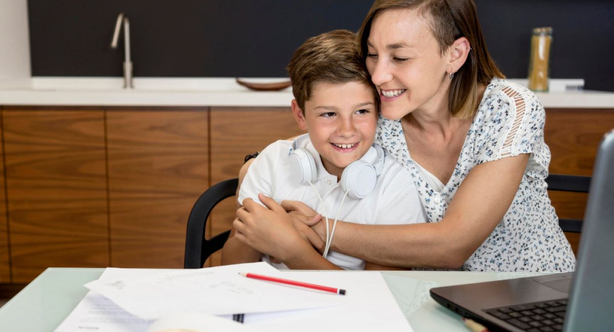 Mother hugging her smiling son while he studies at a table with a laptop and schoolwork.